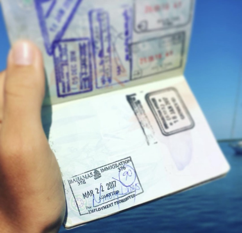 A passport held open over the water, stamped by Bahamas immigration, with a yacht moored behind it