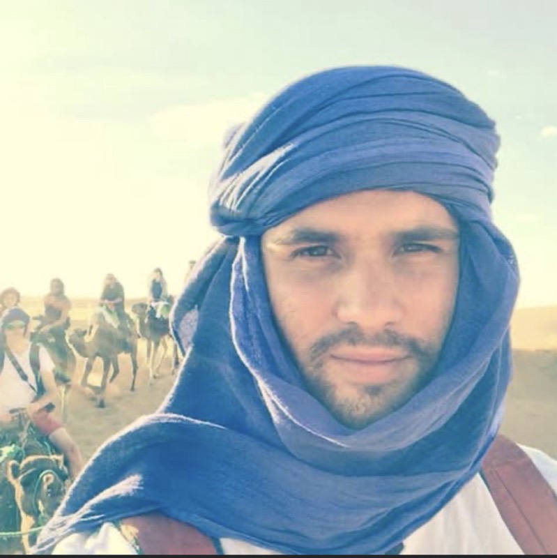 Codie in a blue desert headwrap with a camel train crossing the dunes behind him