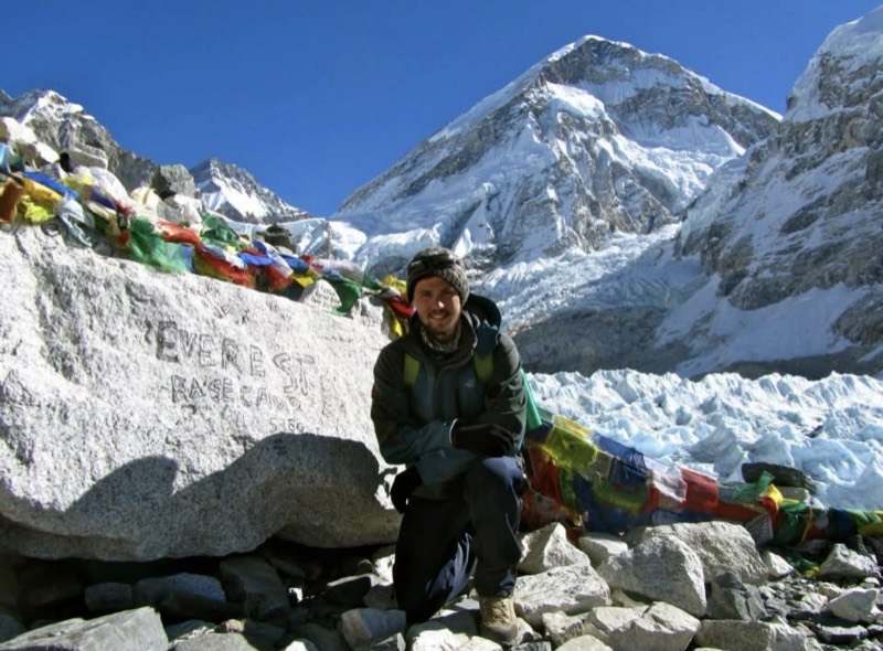Codie kneeling at the Everest Base Camp marker stone, prayer flags and the icefall behind him